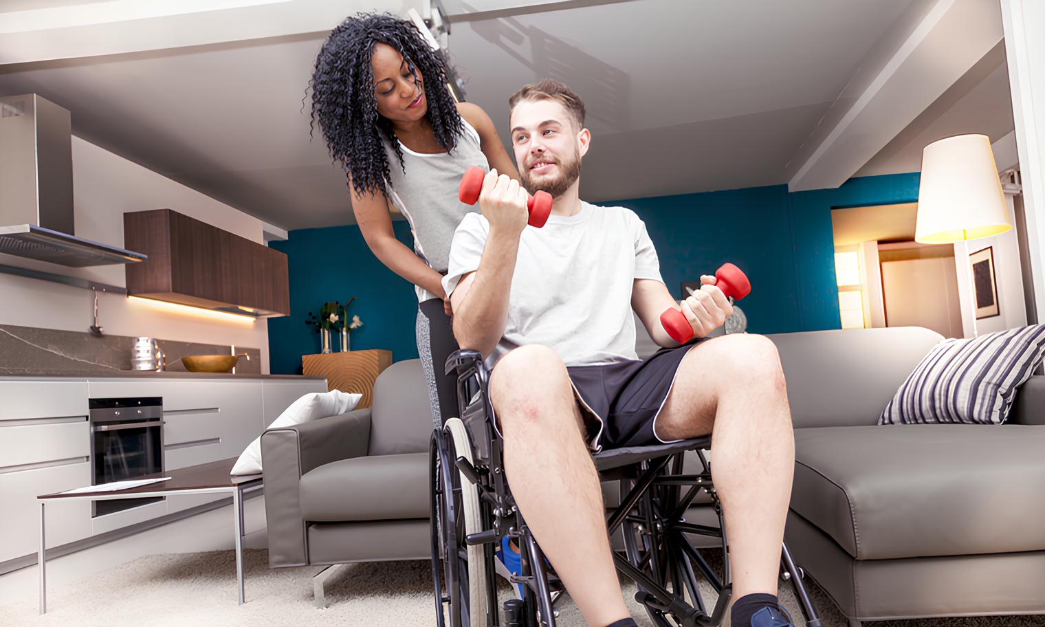 A imagem mostra um homem sorridente em uma cadeira de rodas está fazendo exercícios com halteres vermelhos, enquanto uma mulher com cabelos cacheados escuros o observa por trás, com as mãos em seus ombros. Eles estão em um ambiente interno que parece ser uma sala de estar ou área de convivência com uma cozinha parcialmente visível ao fundo.